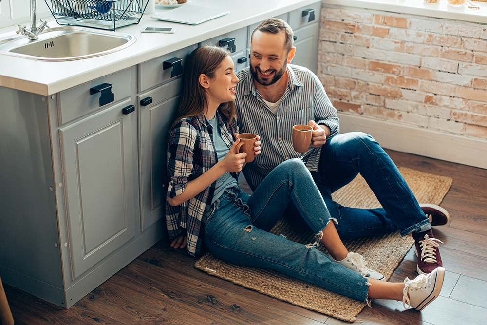couple sitting on kitchen floor smiling and talking while holding coffee mugs